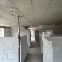 BAR-KAM GROUP SP. Z O.O - Wnętrze surowego domu w trakcie budowy, z widocznymi ścianami z białych bloczków i betonowym stropem podpartym metalową podporą. Widok na korytarz między ścianami.