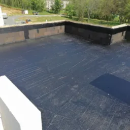 Widok z góry na płaski dach pokryty czarną hydroizolacją, z widocznymi fragmentami muru i zielenią w tle. Detal zakończenia prac hydroizolacyjnych na dachu.