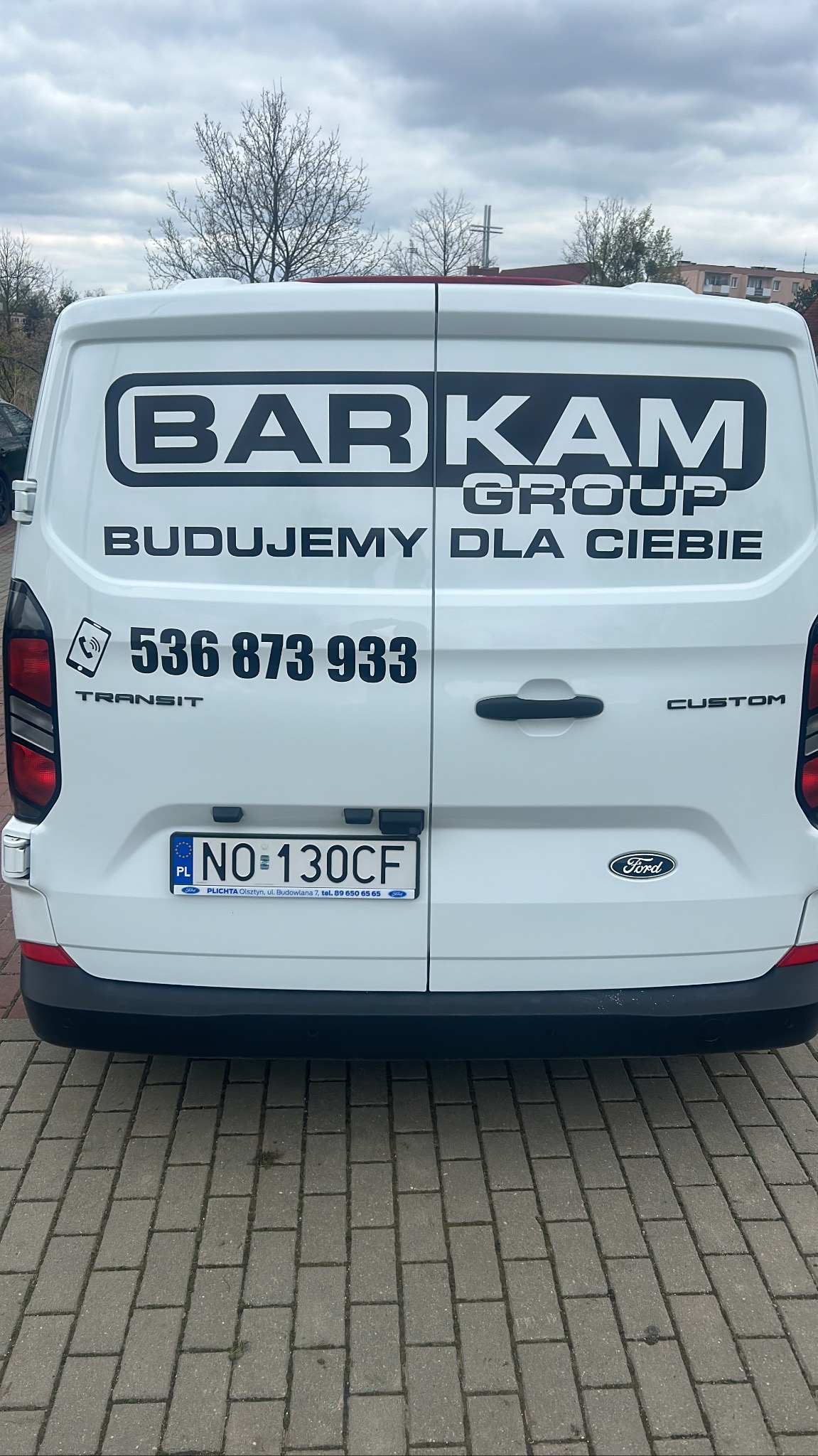 Tył białego samochodu dostawczego Ford Transit Custom z dużym logo firmy budowlanej BARKAM GROUP i numerem telefonu 536 873 933, zaparkowanego na brukowanym chodniku.
