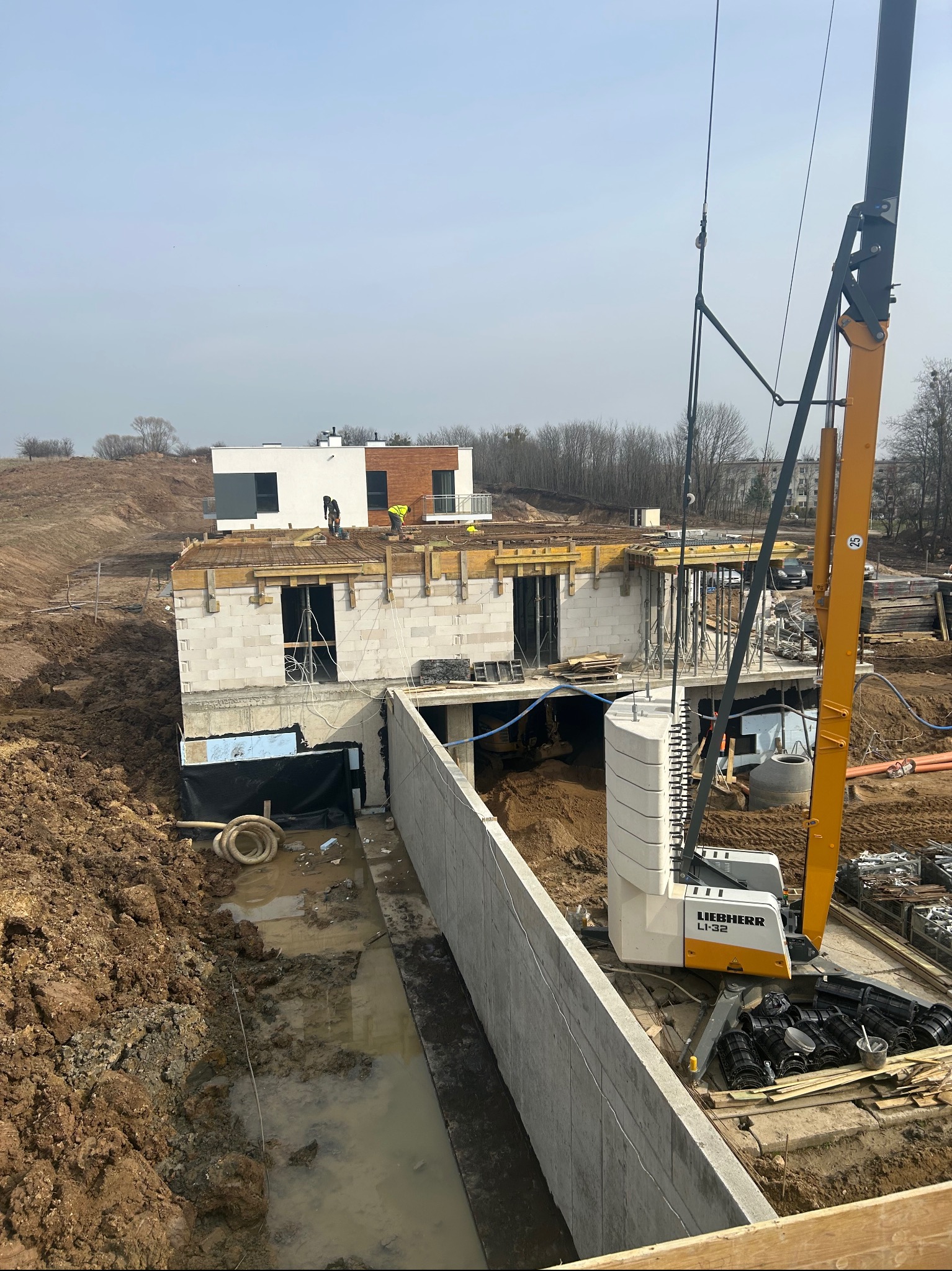 Budowa domu w stanie surowym otwartym z widocznymi fundamentami, ścianami z bloczków, szalunkiem stropu oraz żurawiem budowlanym Liebherr LI-32 na placu budowy.