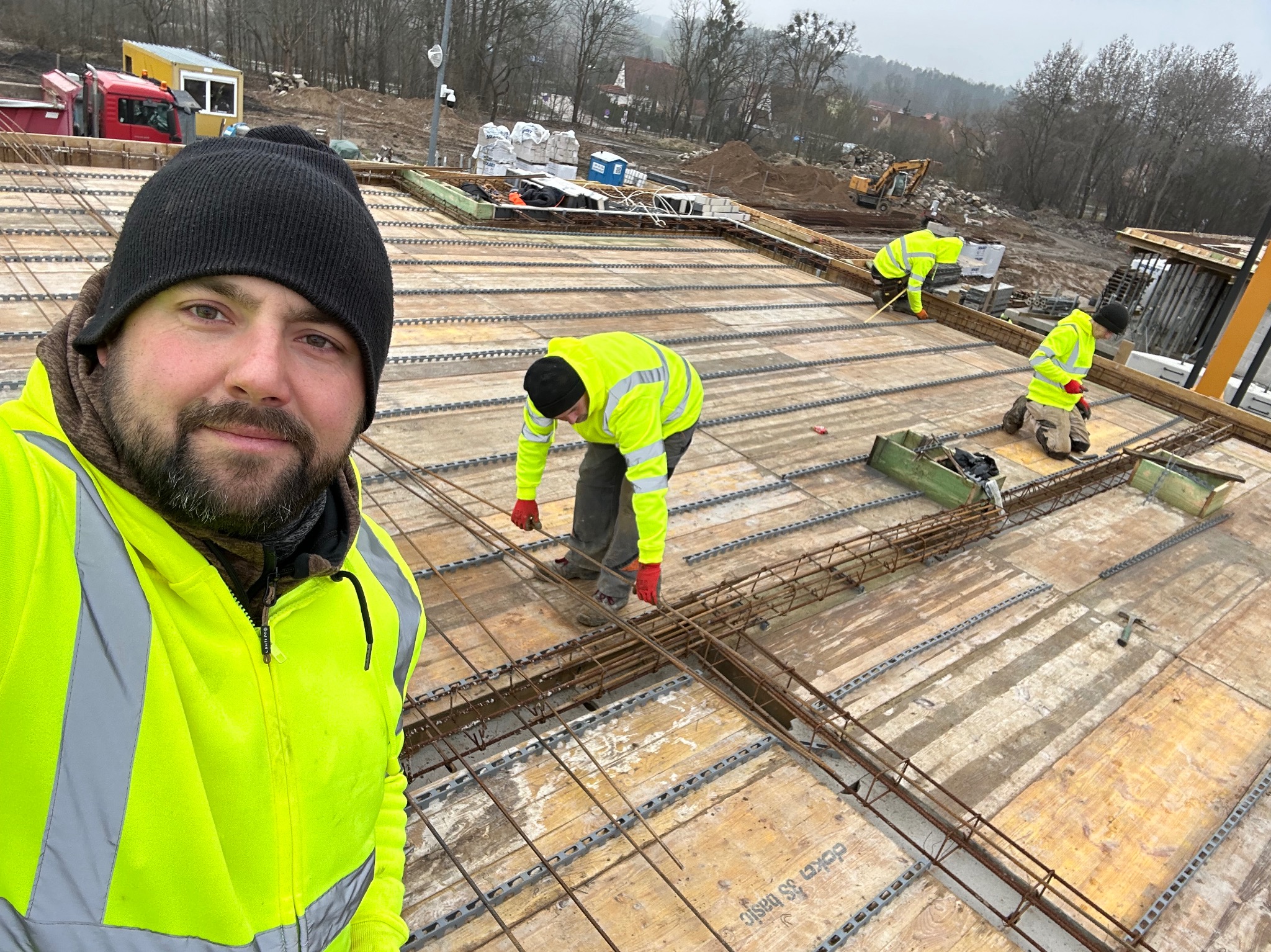 Pracownik w neonowej kurtce robiący selfie na budowie, widoczny zbrojony szkielet konstrukcji, w tle pracują inni robotnicy i stoi ciężarówka.