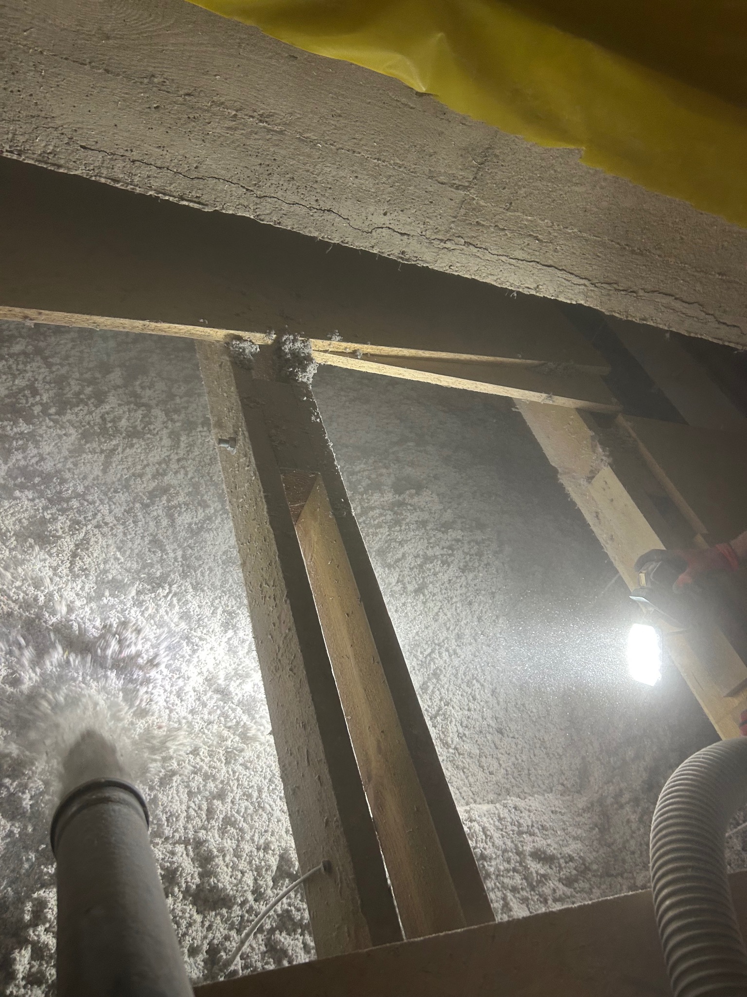 Aplikacja izolacji w przestrzeni poddasza, widoczna dysza rozpylająca materiał izolacyjny, drewniana konstrukcja i oświetlenie robocze. Detal z etapu prac.