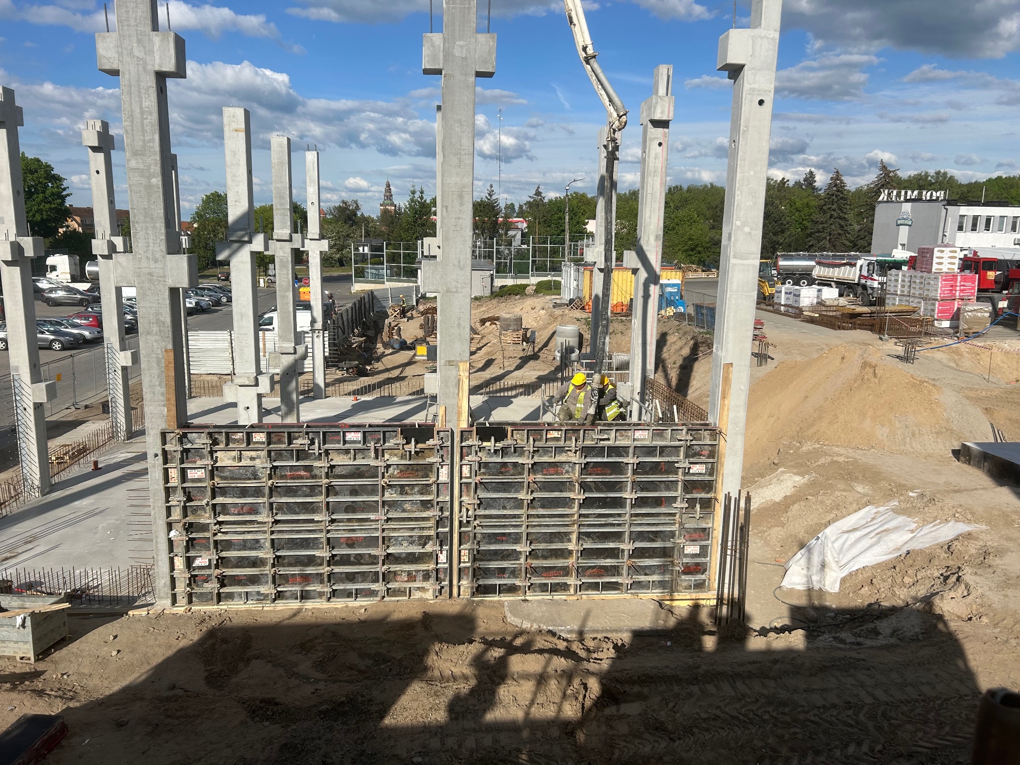 Budowa domu: betonowe słupy konstrukcyjne w szalunkach, widoczni pracownicy w żółtych kaskach, na tle błękitnego nieba z chmurami. Etap wznoszenia budynku.