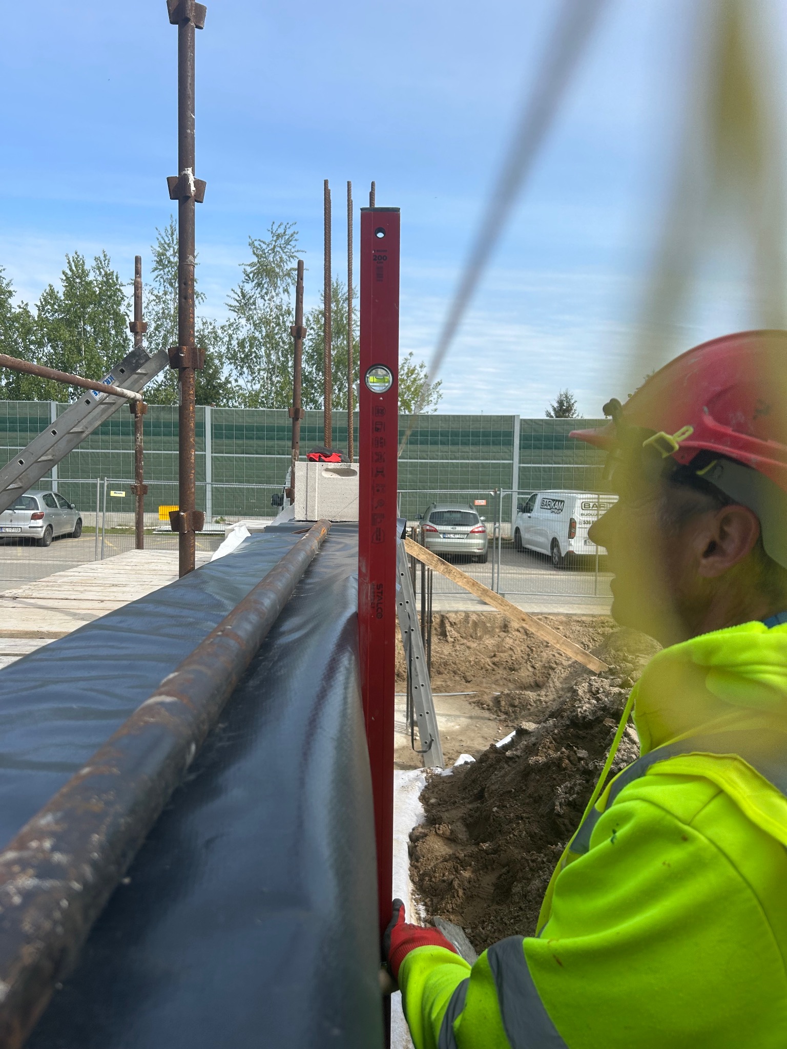 Pracownik w kasku i odblaskowej kamizelce używa poziomicy do sprawdzenia pionu konstrukcji szalunkowej fundamentu na placu budowy. Widoczne są elementy zbrojenia.