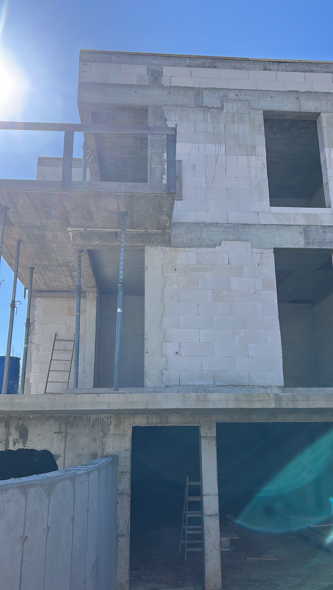 Surowy stan dwukondygnacyjnego domu z betonu komórkowego, widoczne rusztowania i drabiny, błękitne niebo w tle. Budowa w toku, brak wykończenia elewacji.