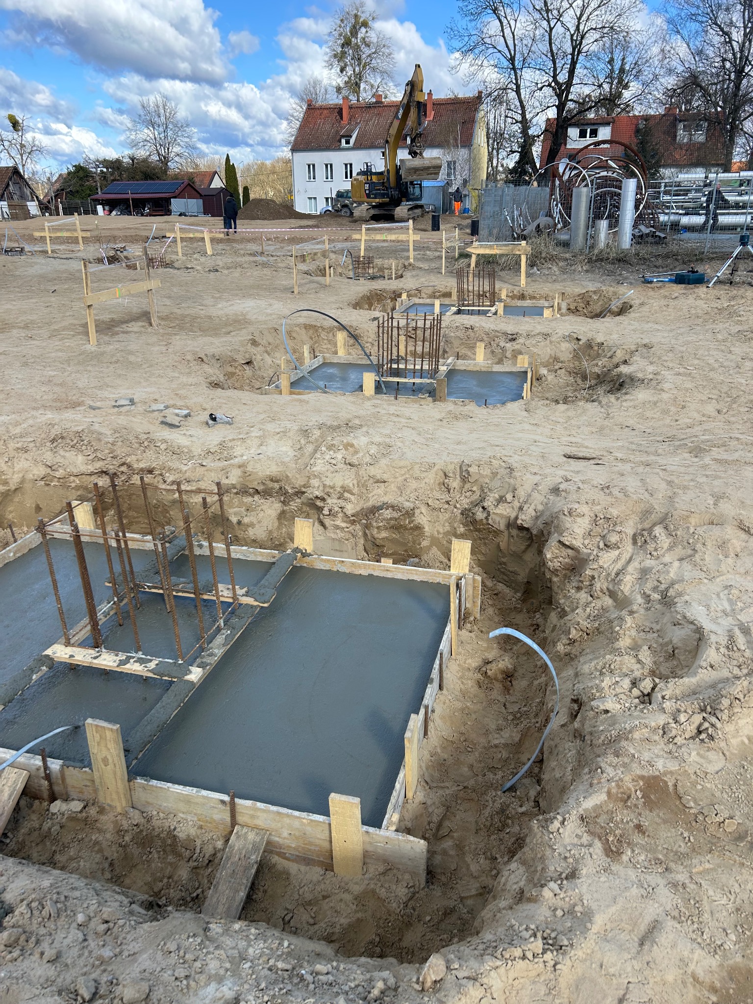 Wylane fundamenty z drewnianym szalunkiem i zbrojeniami na placu budowy. W tle budynki mieszkalne i koparka.