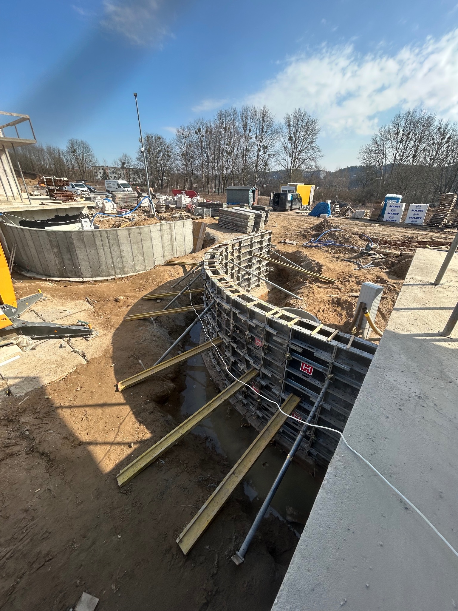 Wykop fundamentów z szalunkiem łukowym, wsparty żółtymi belkami. W tle widoczny częściowo zalany betonem fundament i plac budowy z materiałami.