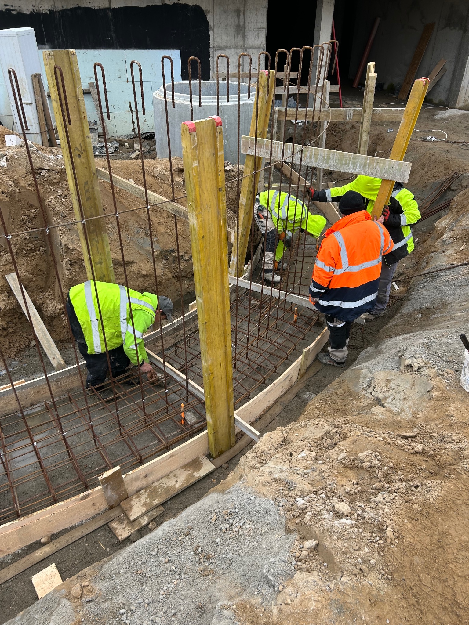 Prace zbrojeniowe fundamentów pod budowę w Lidzbarku Warmińskim. Widoczni pracownicy w odblaskowych ubraniach, drewniane szalunki i stalowe pręty zbrojeniowe.
