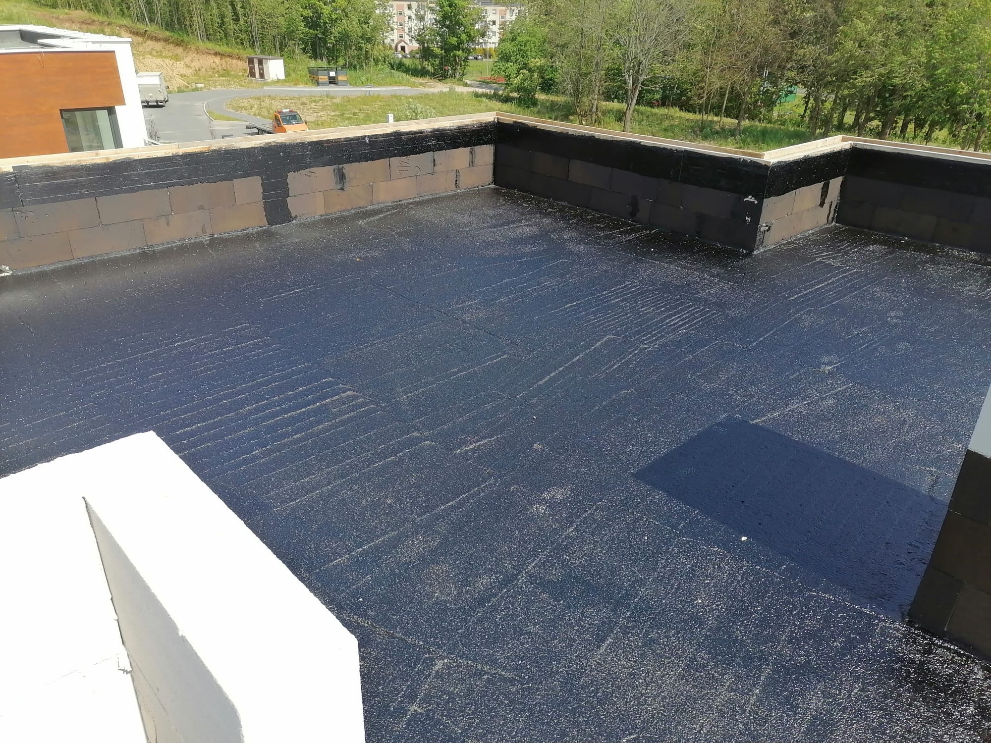 Widok z góry na płaski dach pokryty czarną hydroizolacją, z widocznymi fragmentami muru i zielenią w tle. Detal zakończenia prac hydroizolacyjnych na dachu.