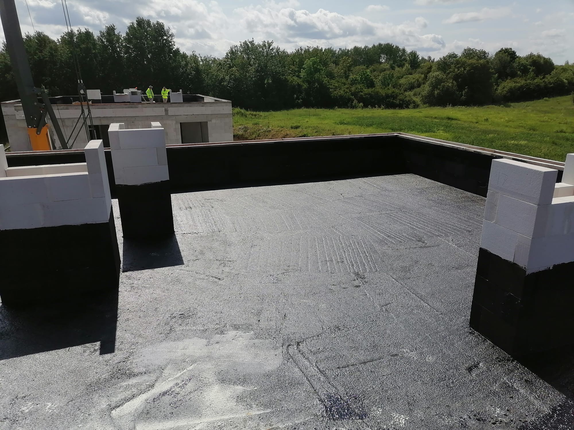 Budowa domu z betonu komórkowego: widok z góry na izolowany fundament i wznoszone ściany. W tle zieleń i pracownicy na kolejnym etapie budowy.