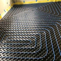 Heatlink Sp z o.o. - Niebieskie rurki ułożone w pętle na czarnej macie z wypustkami, instalacja ogrzewania podłogowego w trakcie montażu, widoczne taśmy zabezpieczające ściany.