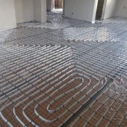 Heatlink Sp z o.o. - Białe rury ogrzewania podłogowego ułożone w pętle na metalizowanej folii, widoczne siatki mocujące, surowy stan wykończenia wnętrza.