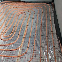Heatlink Sp z o.o. - Pomarańczowe rurki ogrzewania podłogowego zamocowane na srebrnej folii z czerwoną siatką, widoczne czarne mocowania i fragment ściany z wystającymi kablami.