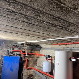 Heatlink Sp z o.o. - Kotłownia z nieocieplonym stropem, widoczne rury grzewcze w izolacji, zbiorniki buforowe i niebieski piec na paliwo stałe.