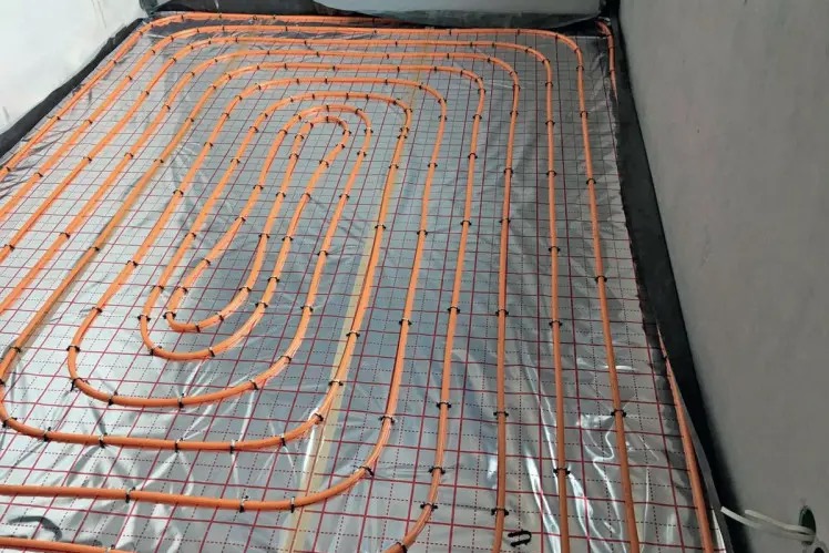 Pomarańczowe rurki ogrzewania podłogowego zamocowane na srebrnej folii z czerwoną siatką, widoczne czarne mocowania i fragment ściany z wystającymi kablami.