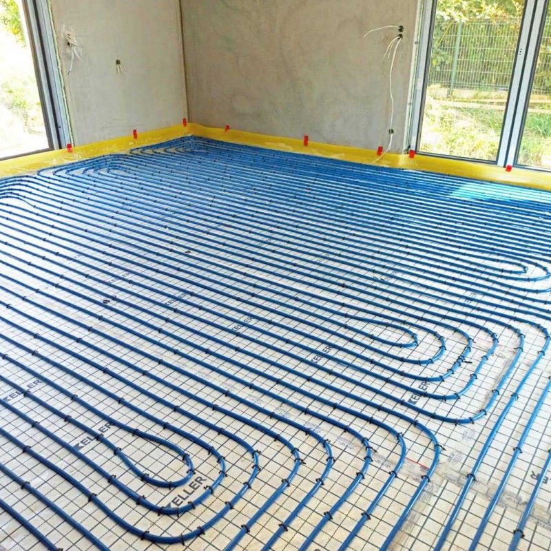 Niebieskie rury ogrzewania podłogowego rozłożone na białej macie z siatką w surowym pomieszczeniu z dużymi oknami, widoczne elementy mocujące i folia ochronna przy ścianach.