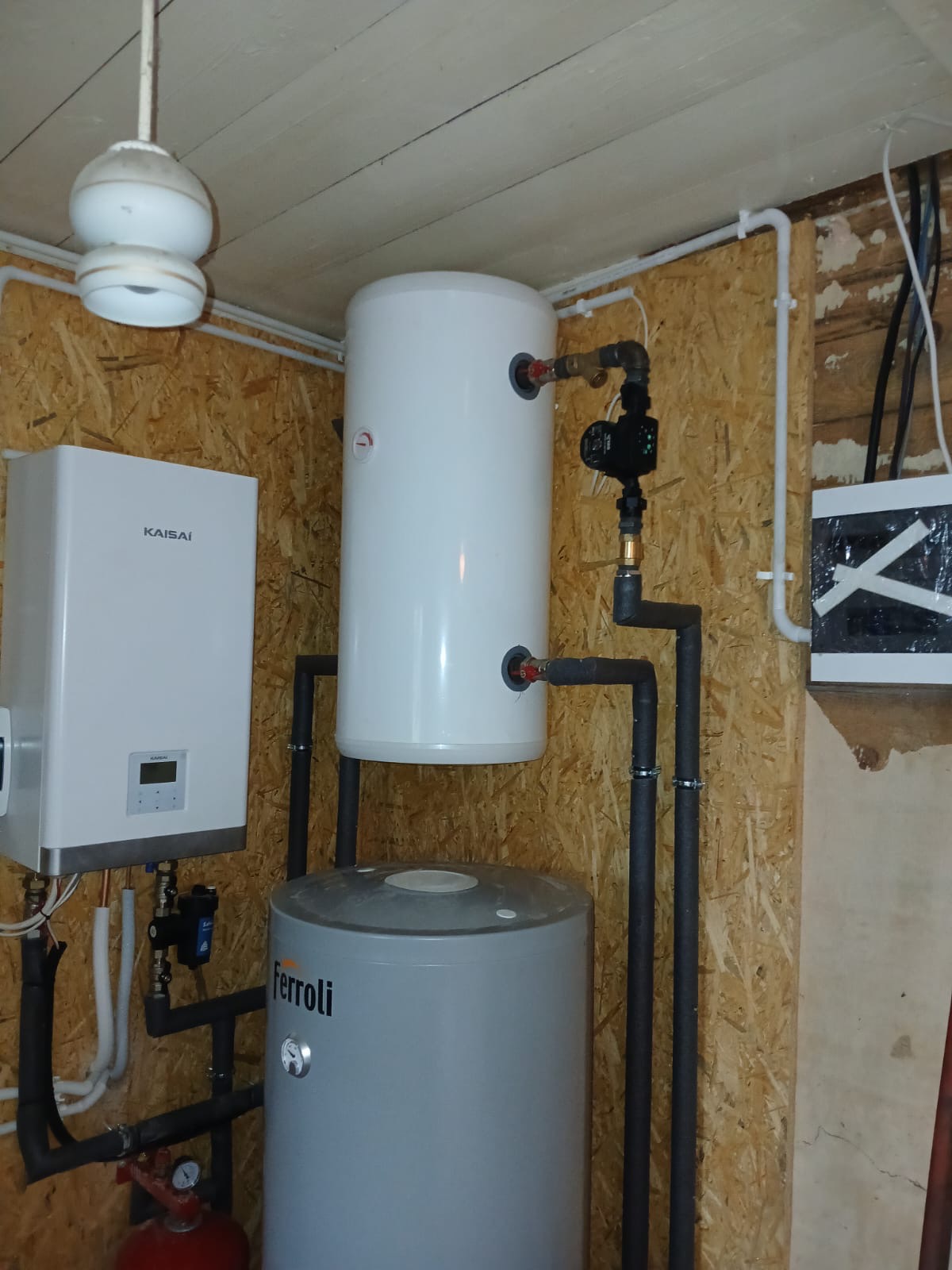Kompaktowa instalacja grzewcza z kotłem Kaisi i zbiornikiem Ferroli, rury w izolacji, widoczne elementy sterujące i zabezpieczające, zamontowana na ścianie z płyty OSB.