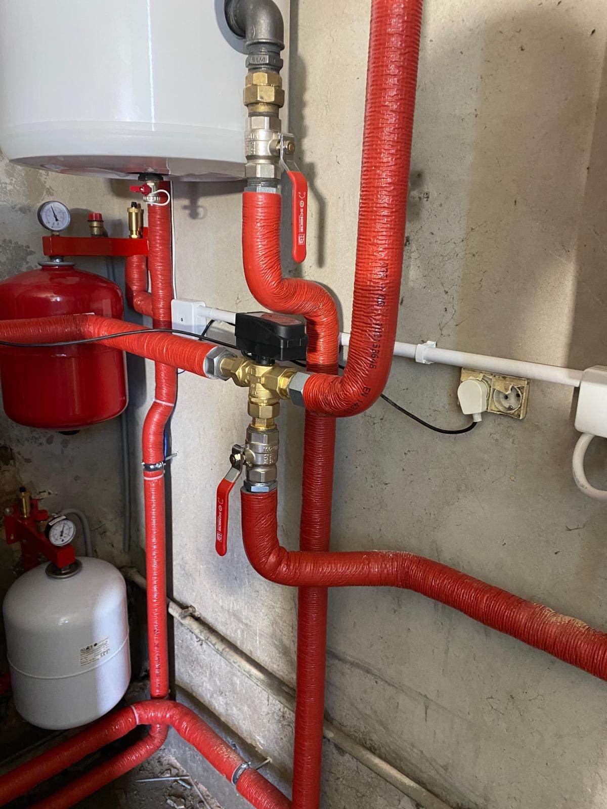 Instalacja centralnego ogrzewania z czerwonymi rurami izolowanymi, manometry, zawory i elementy elektryczne na tle surowej ściany. Widoczny biały zbiornik wyrównawczy i naczynie przeponowe.