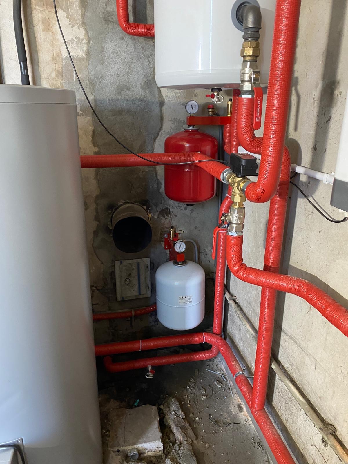 Instalacja centralnego ogrzewania w piwnicy: widoczny zbiornik wyrównawczy, rury w czerwonej izolacji termicznej, manometry i zawory.