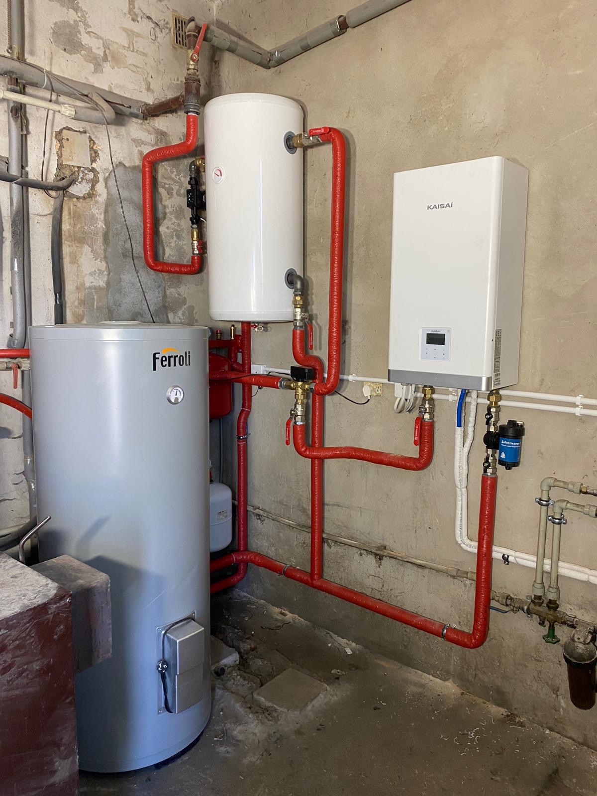 Zestaw urządzeń grzewczych: pionowy szary zasobnik Ferroli z termometrem, biały zasobnik i biały piec Kaisai, połączone rurami w czerwonej izolacji, zamontowane na szarej, surowej ścianie.