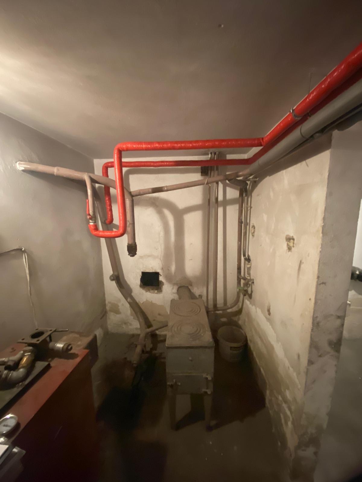 Stara kotłownia z piecem na paliwo stałe, widoczne rury grzewcze w kolorze czerwonym i szarym, ściany z ubytkami tynku, podłoga prawdopodobnie zalana wodą, w tle wiadro.