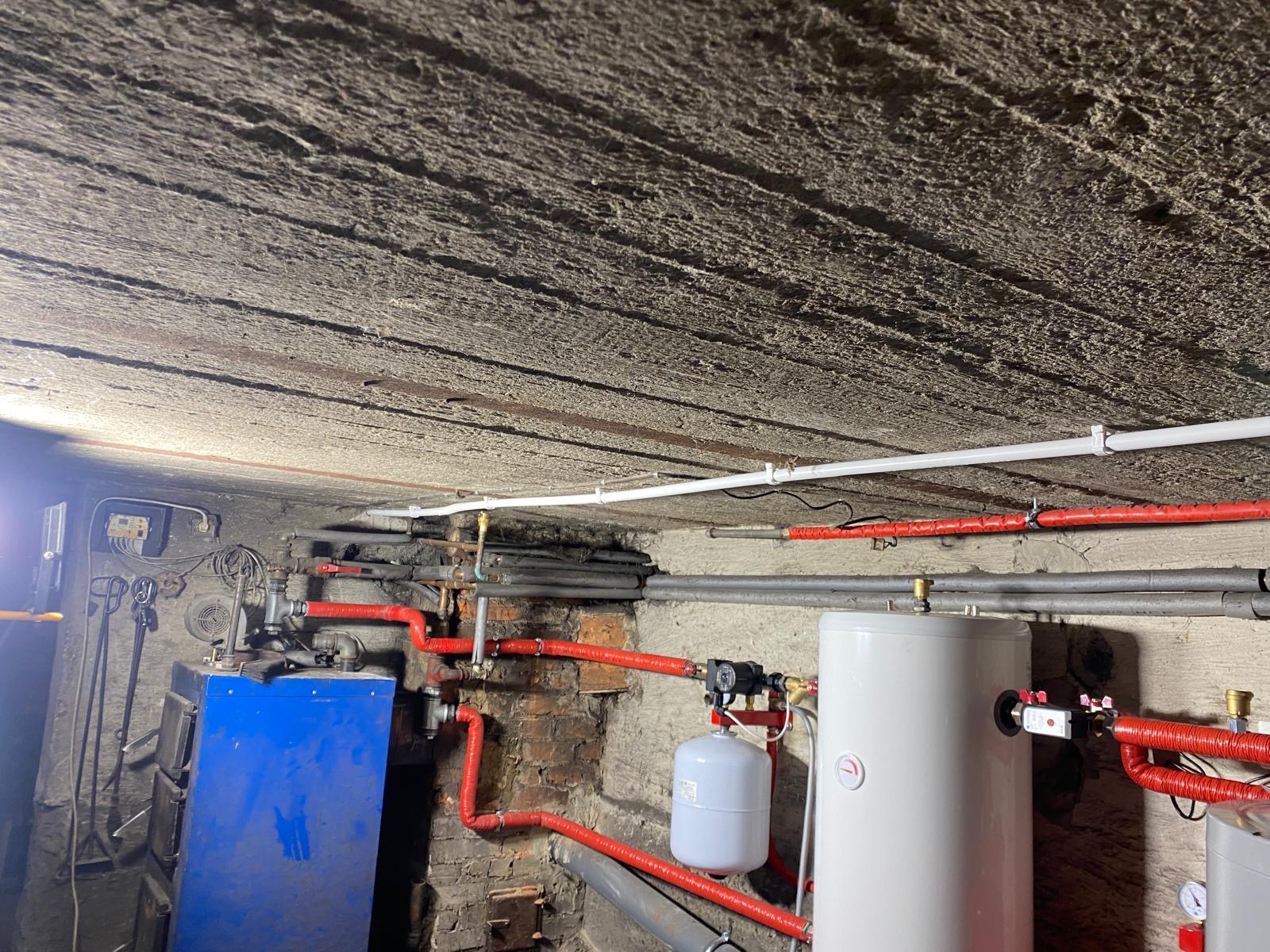 Kotłownia z nieocieplonym stropem, widoczne rury grzewcze w izolacji, zbiorniki buforowe i niebieski piec na paliwo stałe.