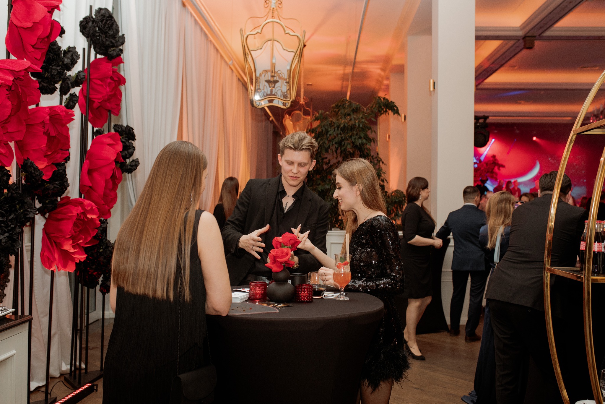 Iluzjonista w czarnym garniturze prezentuje trik z czerwonymi różami dwóm kobietom przy okrągłym stoliku na tle eleganckiego przyjęcia z dekoracjami z czerwonych i czarnych kwiatów.