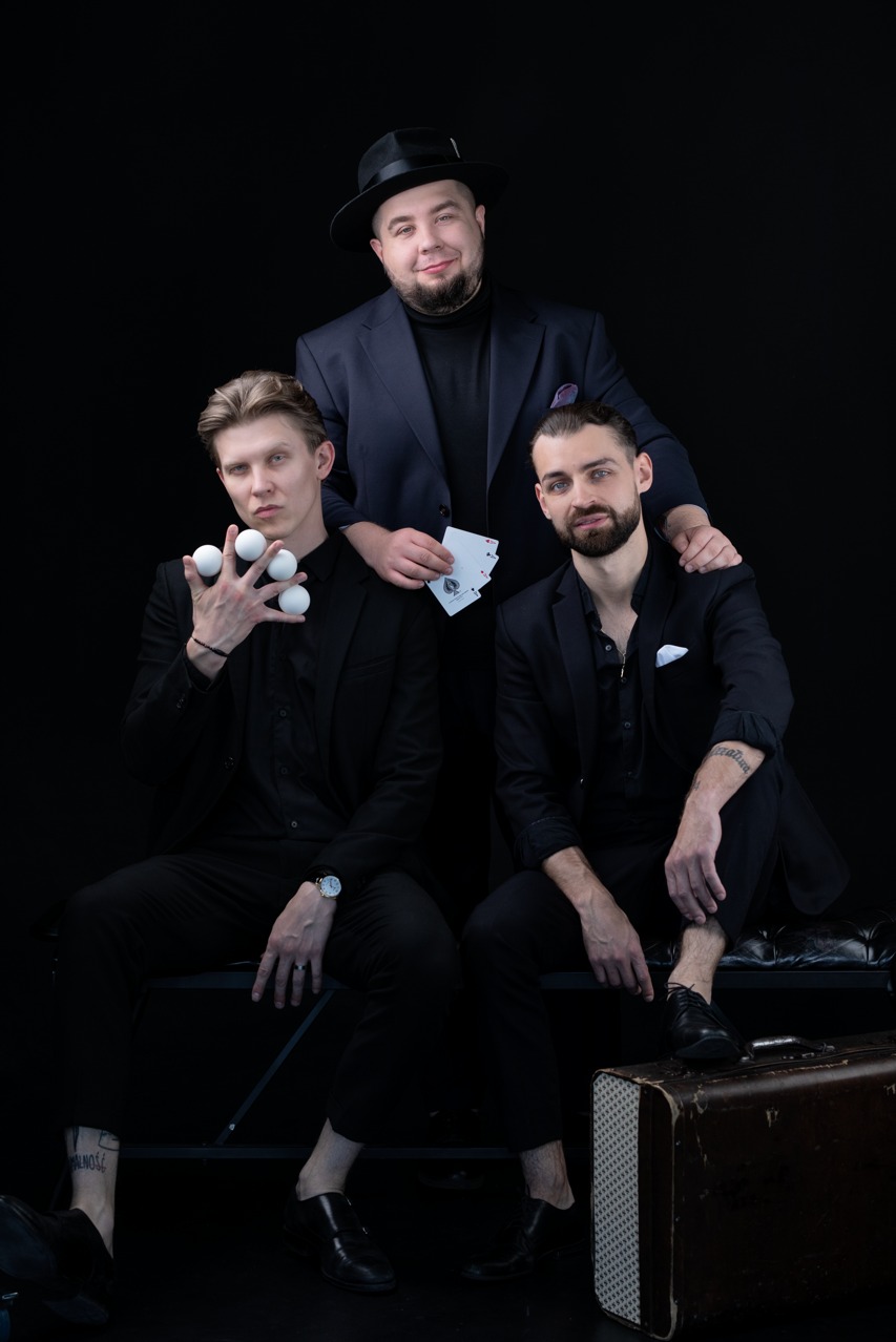 Trzech mężczyzn w czarnych garniturach pozuje na czarnym tle: jeden w kapeluszu trzyma karty, drugi żongluje białymi kulkami, a trzeci opiera się o jego ramię, obok leży stara walizka.
