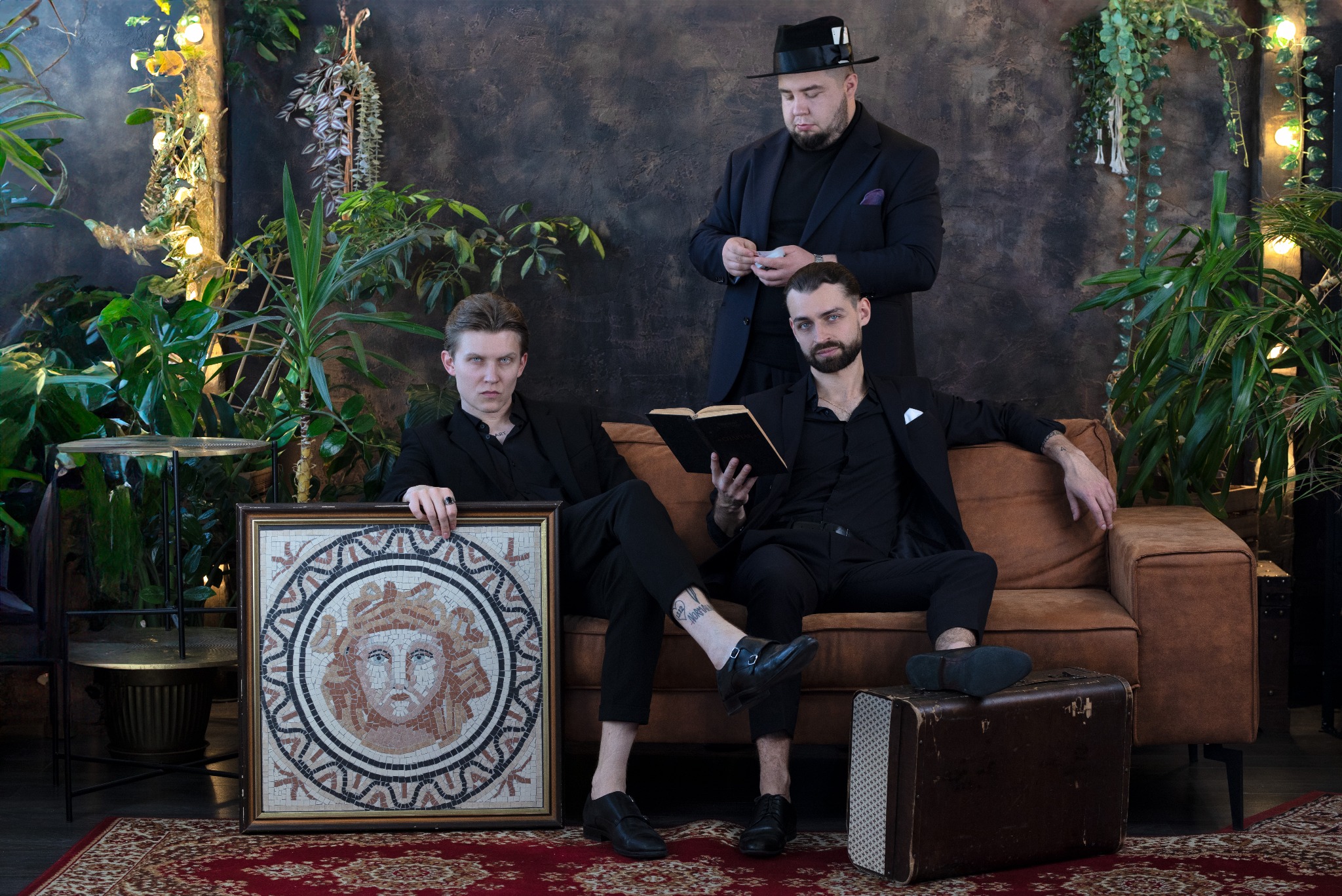 Trzech elegancko ubranych mężczyzn w Poznaniu pozuje w otoczeniu roślin: jeden w kapeluszu trzyma karty, drugi czyta książkę, a trzeci trzyma mozaikę z wizerunkiem twarzy.