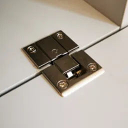 Metalowy zawias meblowy zamontowany na białej płycie meblowej, widoczne śruby mocujące.