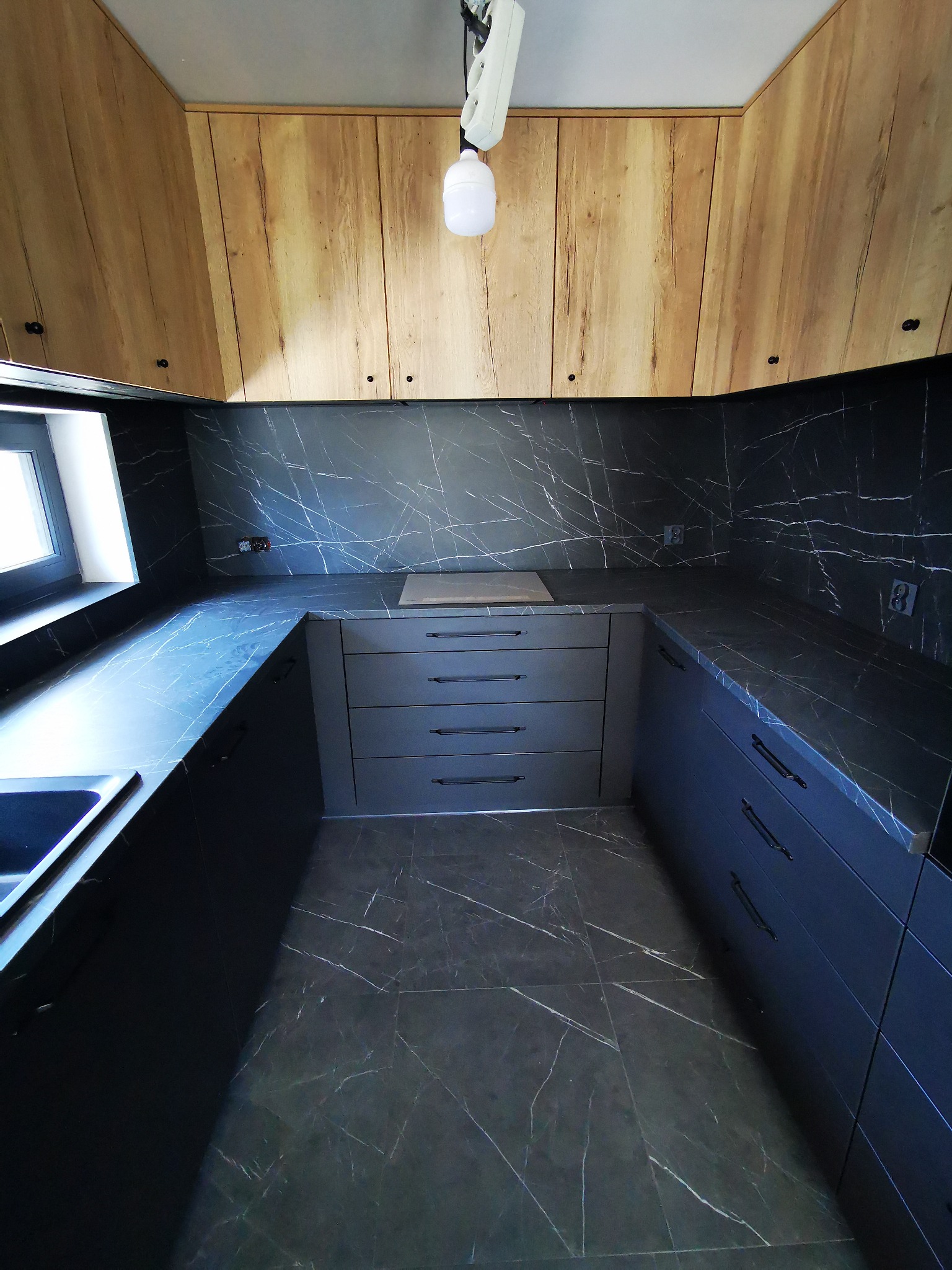 Nowoczesna kuchnia z czarnymi marmurkowymi blatami i frontami szafek, drewniane szafki wiszące z widocznym oświetleniem roboczym.