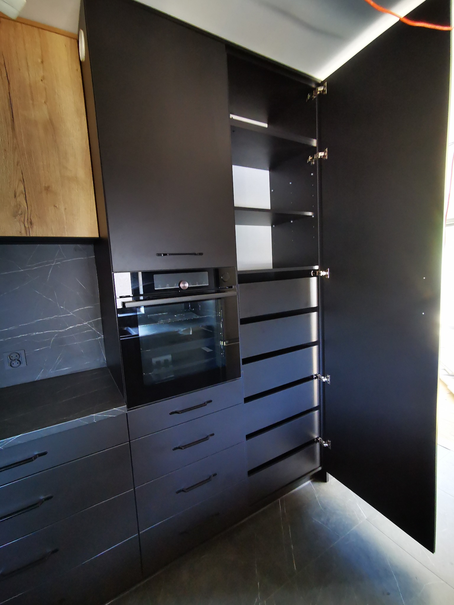 Ciemna zabudowa kuchenna z piekarnikiem i szafkami, otwarte drzwi szafy z półkami i szufladami, marmurowy blat i ściana.