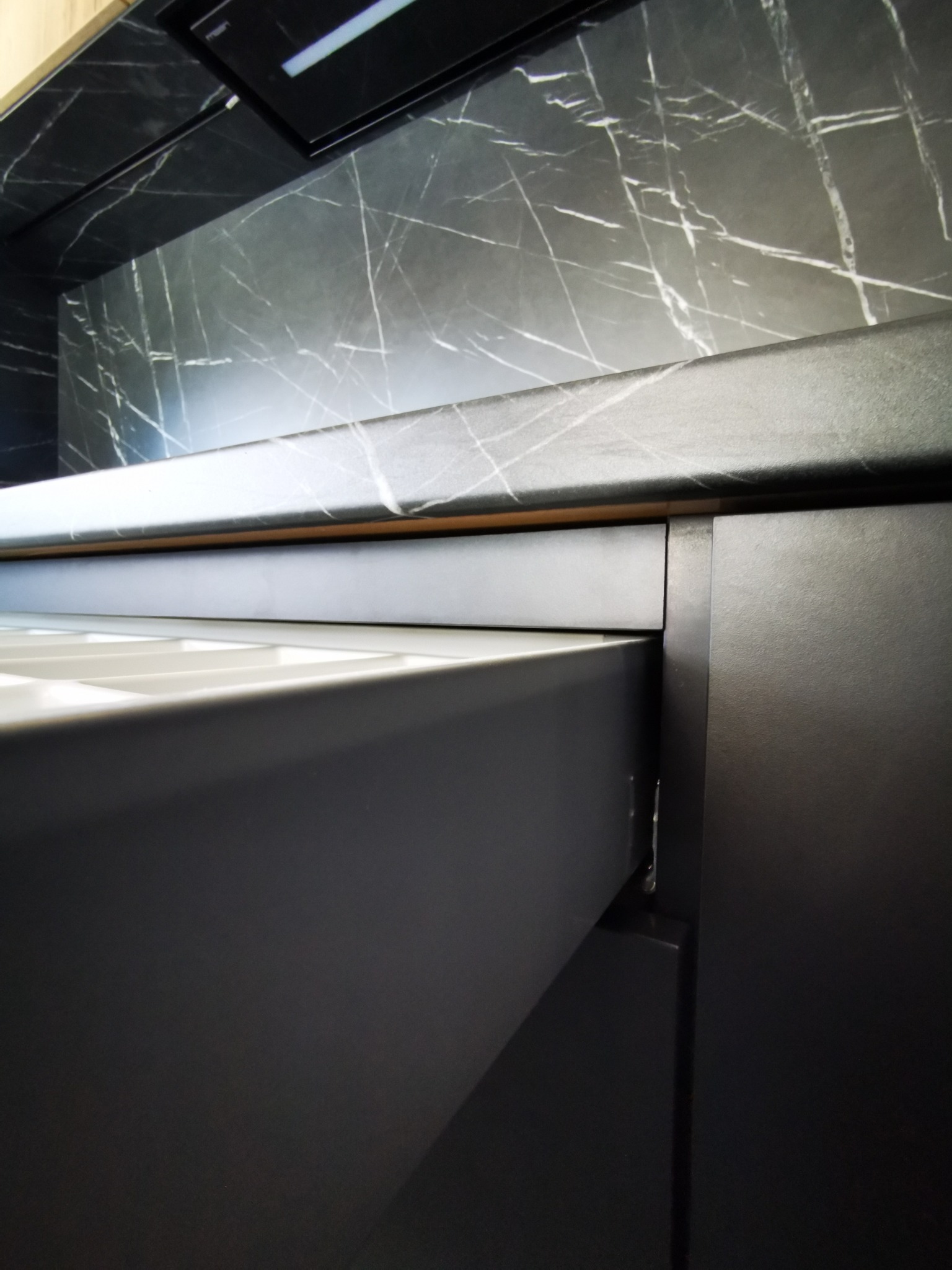 Szczegółowe ujęcie wysuwanej szuflady z organizerem w nowoczesnej, czarnej kuchni z blatem i ścianą w marmurkowy wzór.