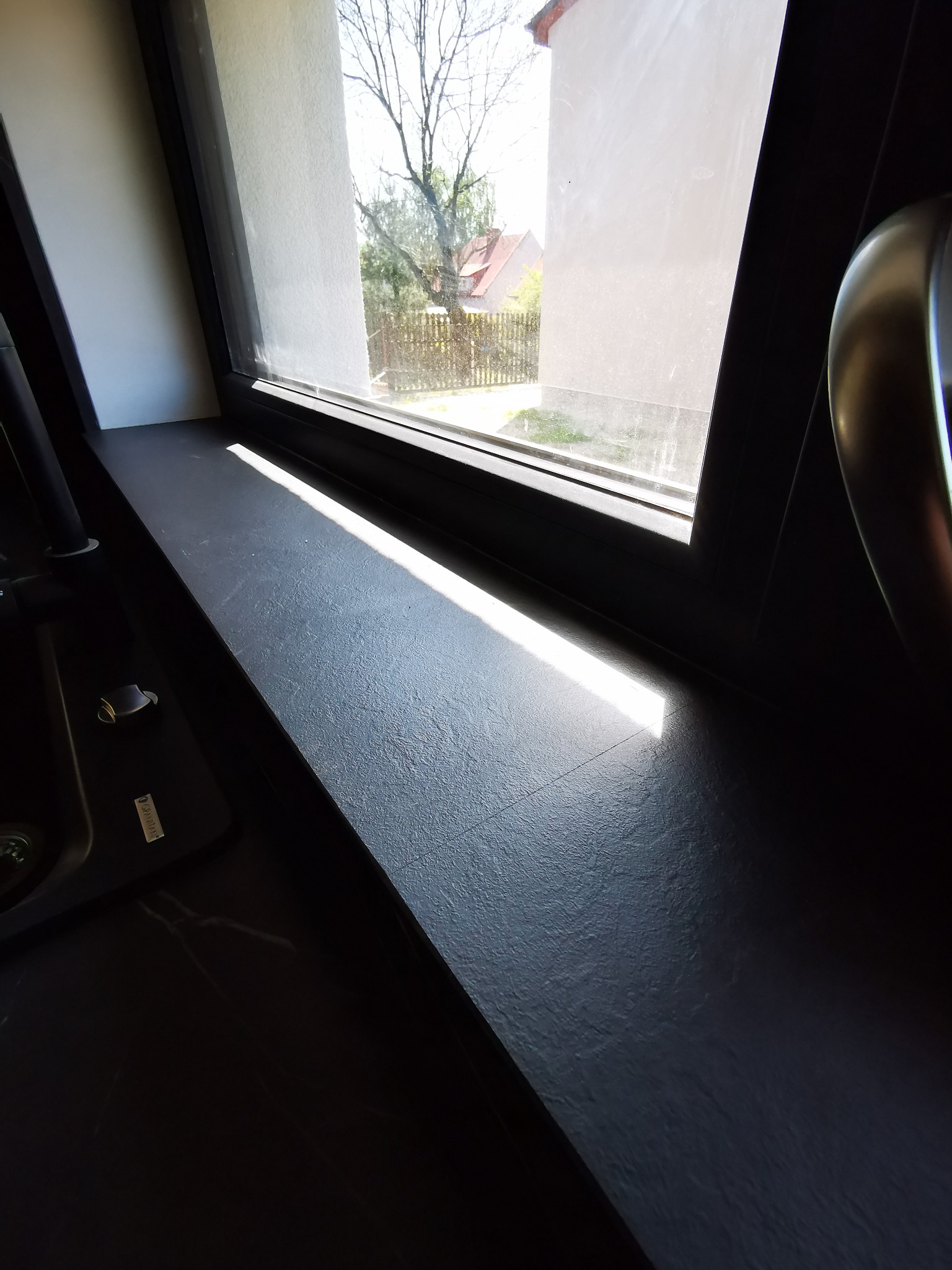 Czarny, teksturowany blat pod oknem w kuchni, z widocznym fragmentem zlewu i elementami armatury, oświetlony promieniami słońca.