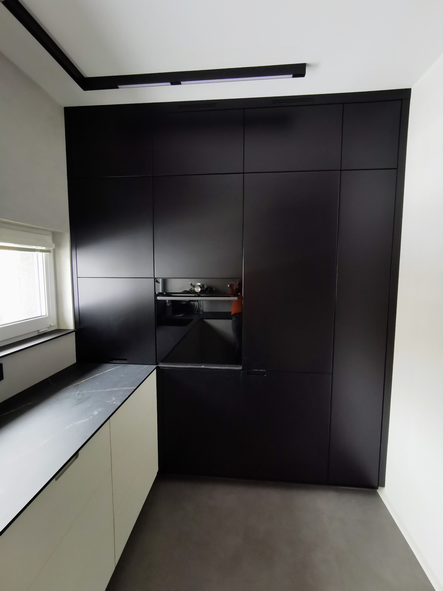 Nowoczesna kuchnia: zabudowa na całej ścianie z czarnymi, matowymi frontami, piekarnik wkomponowany w szafę, białe dolne szafki z blatem imitującym marmur, szara podłoga.
