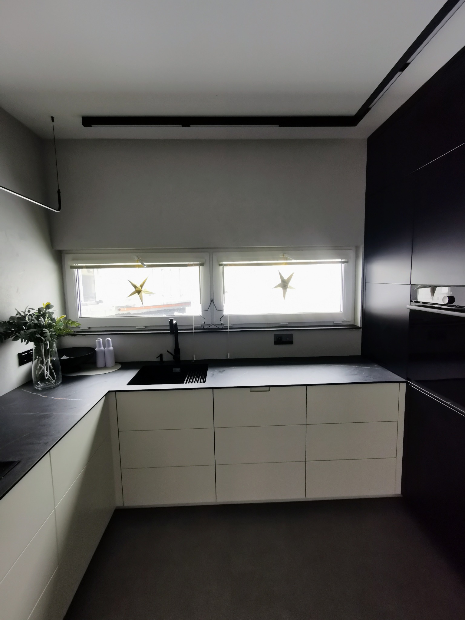 Nowoczesna kuchnia z białymi szafkami dolnymi, czarnym blatem i czarnym zlewozmywakiem, oświetlenie szynowe na suficie, dekoracje okienne w kształcie gwiazd.