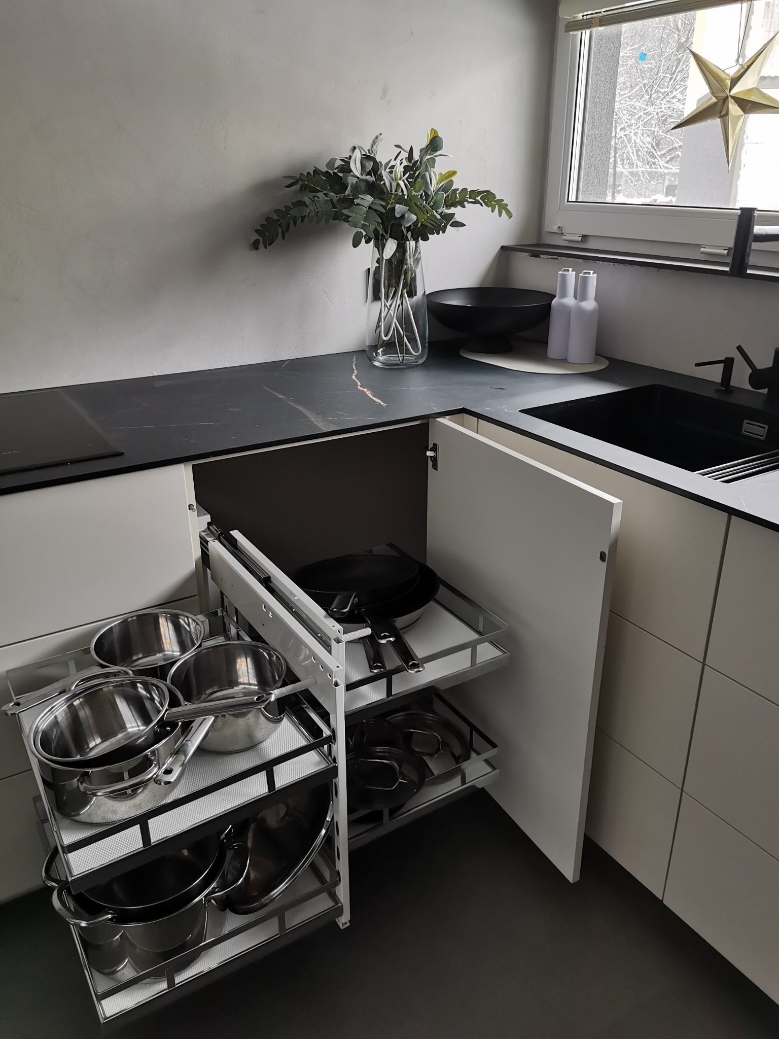 Nowoczesna kuchnia z wysuwanym systemem przechowywania garnków i patelni, widoczne stalowe naczynia na półkach, czarny blat i zlewozmywak, dekoracje okienne w postaci gwiazdy.