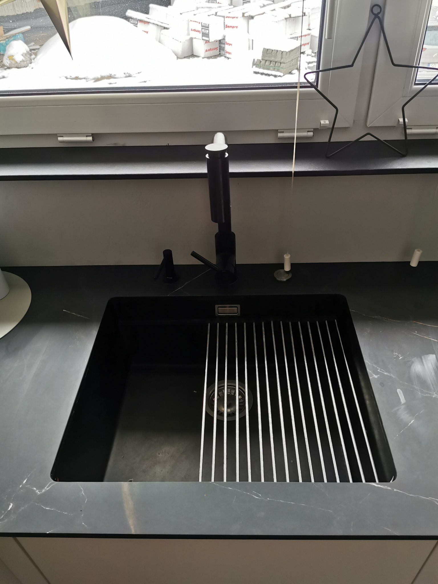 Czarny zlew z ociekaczem i czarną armaturą na blacie imitującym marmur, w tle okno z widokiem na zaśnieżone otoczenie i dekoracja w kształcie gwiazdy.