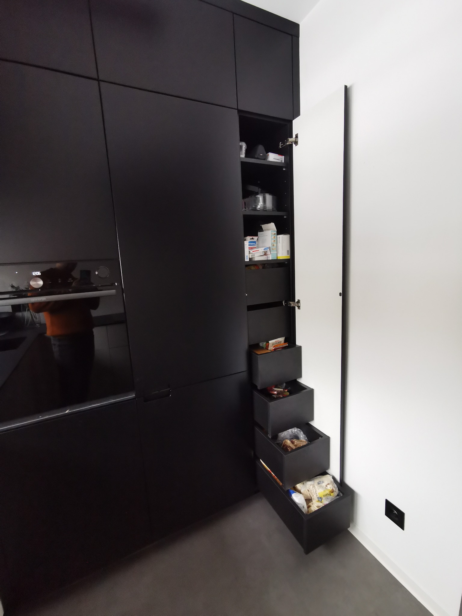 Nowoczesna, czarna zabudowa kuchenna z otwartymi szufladami typu cargo, ukazującymi zawartość spiżarni.