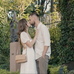 PromoFilm ART - Zakochana para całuje się w parku na tle rzeźb i drzew porośniętych bluszczem, w słoneczny dzień.