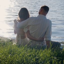 PromoFilm ART - Para siedzi na brzegu jeziora, obejmując się, z widokiem na lśniące słońce odbijające się w wodzie, otoczona zieloną trawą.