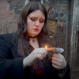 PromoFilm ART - Młoda kobieta w czarnym płaszczu podpala banknoty zapalniczką na tle ceglanego budynku.