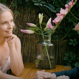 PromoFilm ART - Panna młoda w koronkowej sukni spogląda z uśmiechem na pana młodego podczas kameralnego przyjęcia weselnego, z bukietem lilii i mieczyków w szklanym wazonie na drewnianym stole.