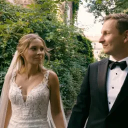 Para młoda spacerująca w parku, panna młoda w koronkowej sukni z welonem, pan młody w smokingu z muchą, w tle zieleń i ceglany mur.