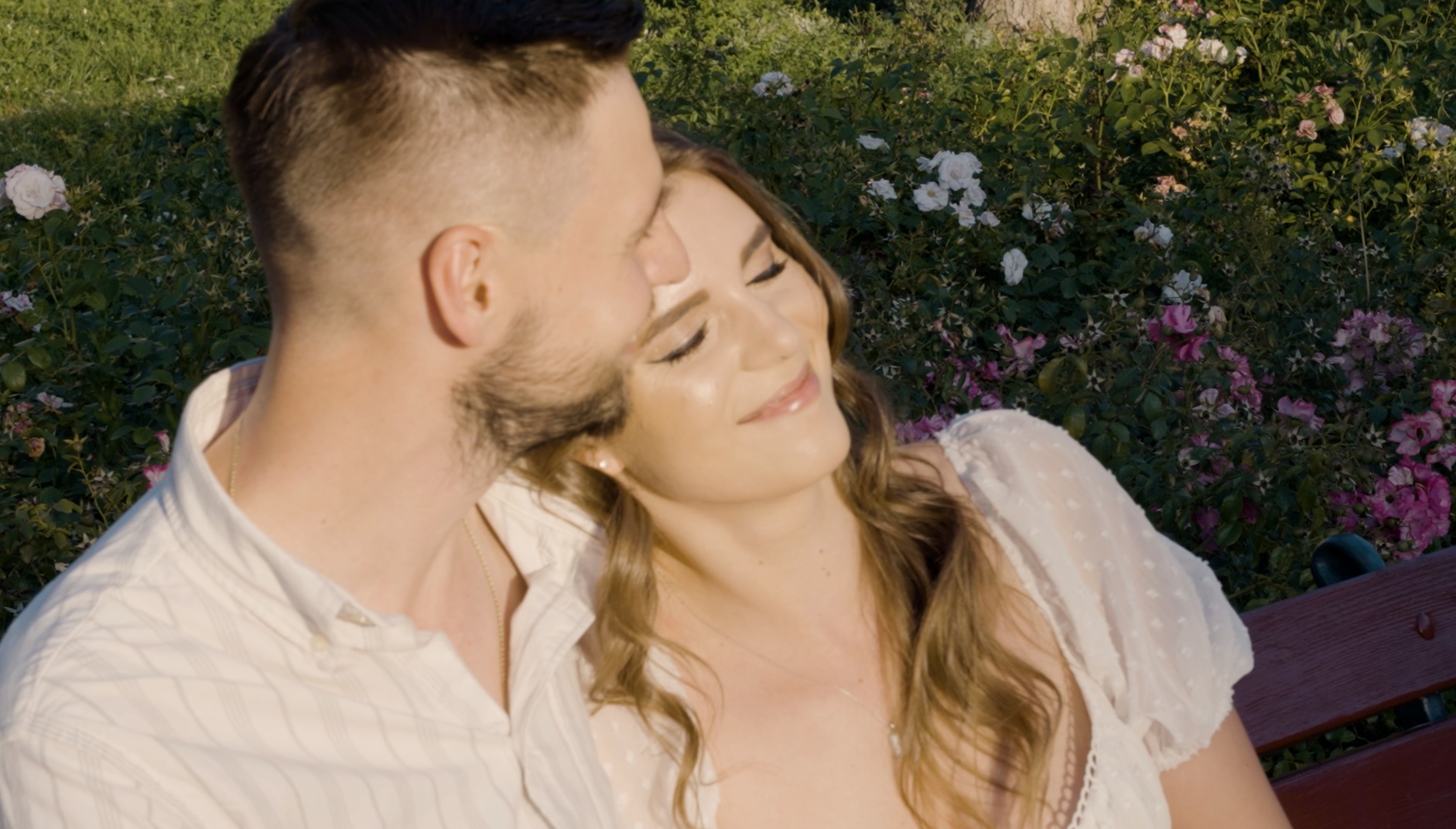 Uśmiechnięta kobieta opiera głowę na ramieniu mężczyzny na tle kwitnących róż w parku, uchwycona w ciepłym, naturalnym świetle.