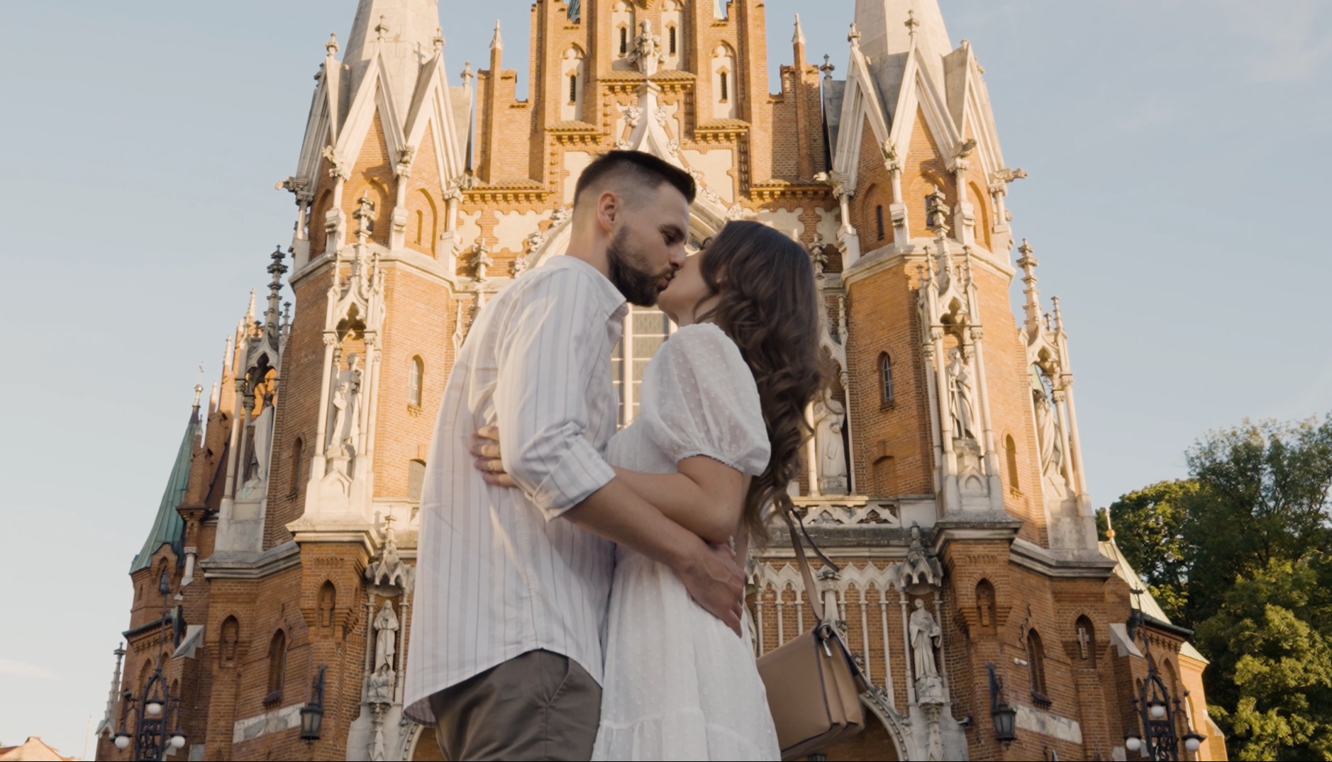 Para całująca się na tle ceglastej, gotyckiej architektury kościoła z licznymi wieżyczkami i zdobieniami.