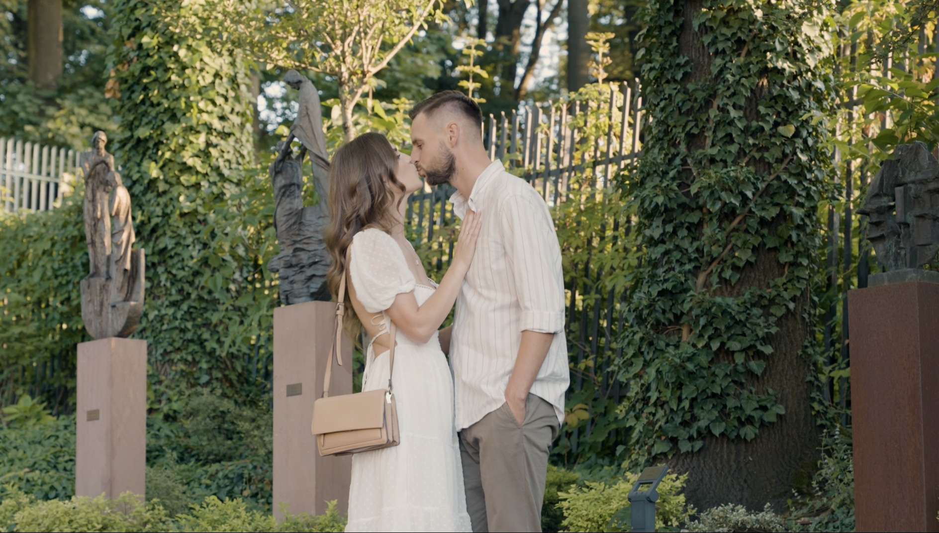 Zakochana para całuje się w parku na tle rzeźb i drzew porośniętych bluszczem, w słoneczny dzień.