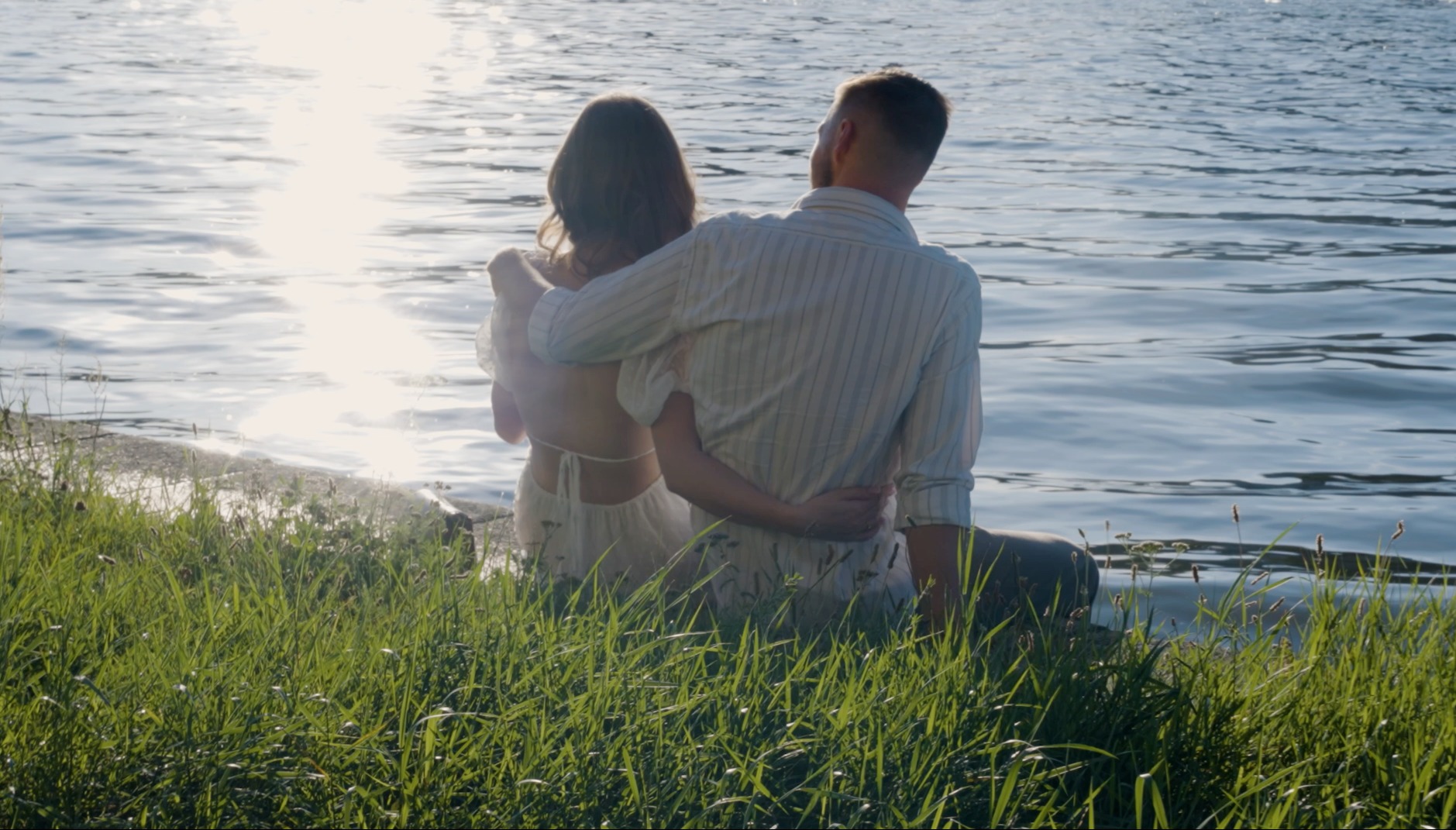 Para siedzi na brzegu jeziora, obejmując się, z widokiem na lśniące słońce odbijające się w wodzie, otoczona zieloną trawą.