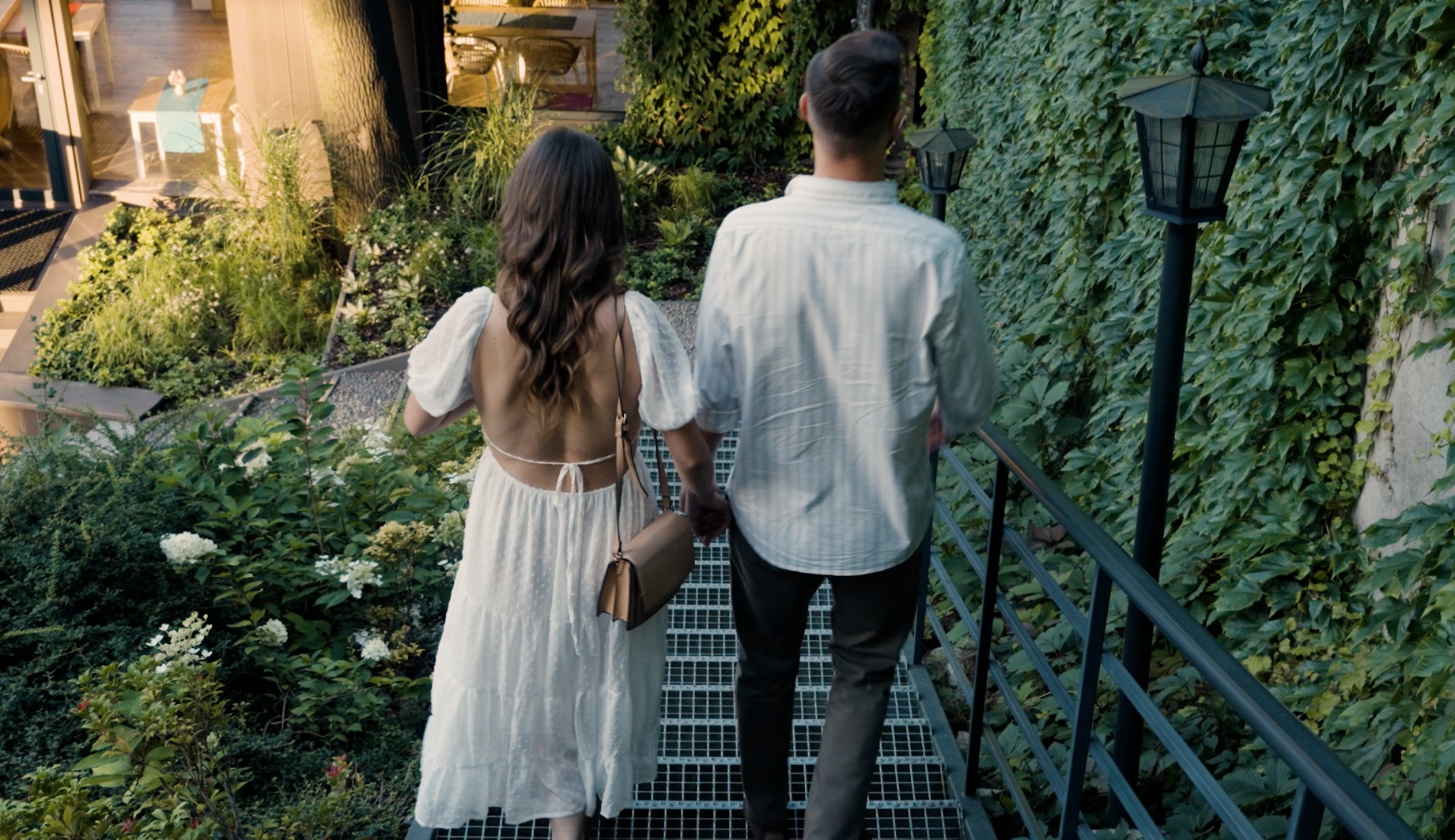 Para młoda schodząca po metalowych schodach otoczonych zielenią, widok z tyłu, kobieta w białej sukience z odkrytymi plecami, mężczyzna w koszuli w paski.