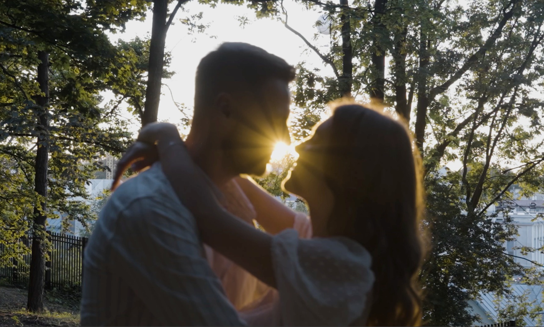 Para zakochanych w objęciach, sylwetki na tle zachodzącego słońca w parku, promienie słoneczne rozświetlają twarze.